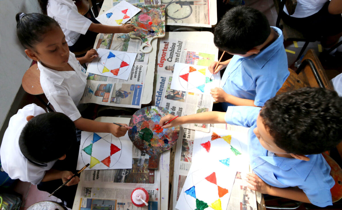 Children seated at school desks drawing colorful shapes.