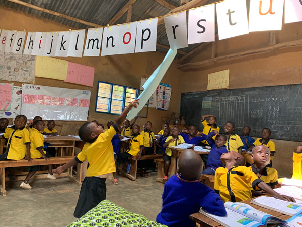 A small child stands in the middle of a classroom, using a long rolled up paper to tap the letter "r" hanging from the ceiling on a long string. The alphabet from the letter "e" to the letter "us" is visible within the frame of the photo.