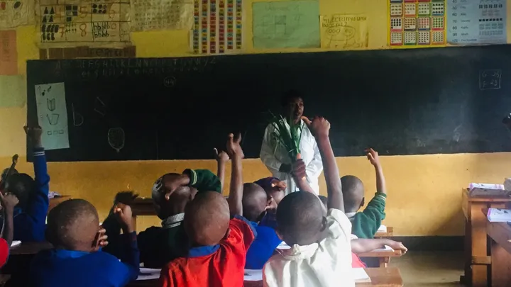 A teacher stands in front of a blackboard, facing a classroom of young students with their hands raised.