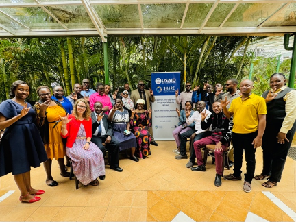 Two rows of people, seated in front and standing in back, At the center is a sign with the logos for the U.S. Agency for International Development and Supporting Holistic and Actionable Research in Education (SHARE). Some of the people are holding their hands in the "I love you" handshape used in some sign languages.