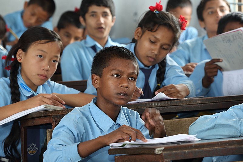 Schoolchildren in blue shirts are seated at shared tables. All of them are holding books.