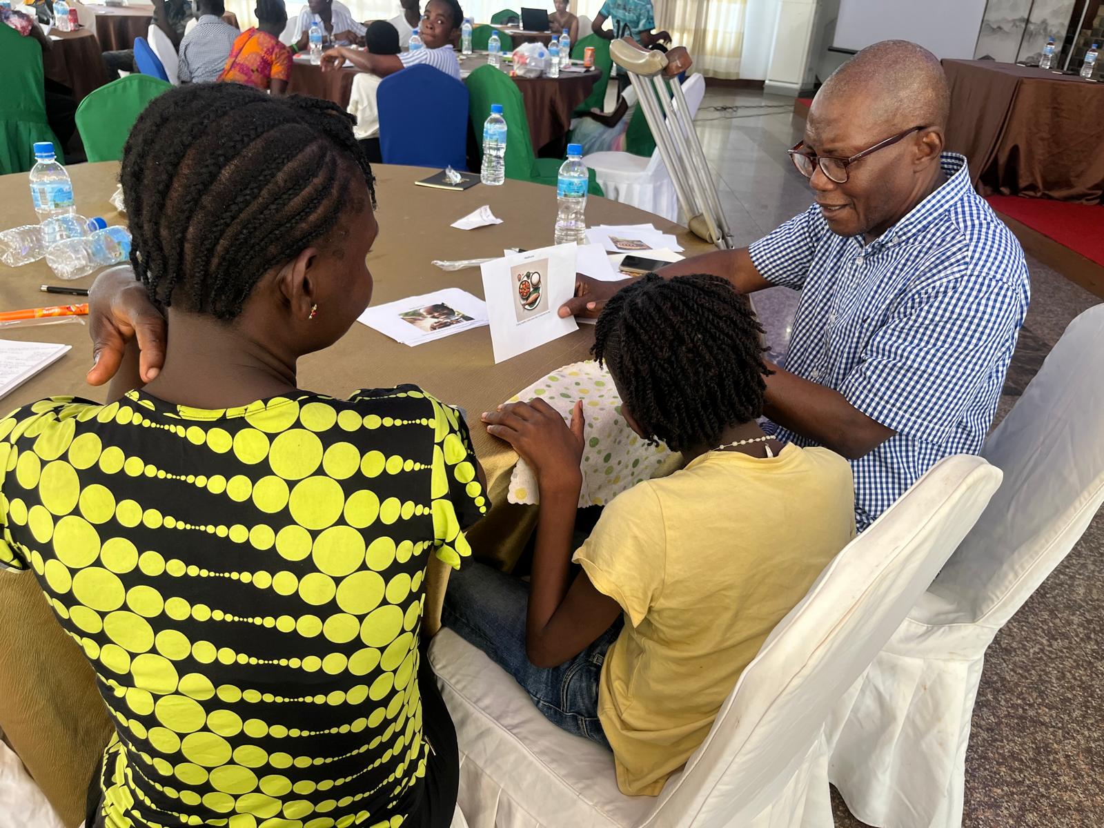 Two adults are seated at a round child with a child seated in between. A man is showing a picture to the child. Other adults and children are visible in the background at other tables.