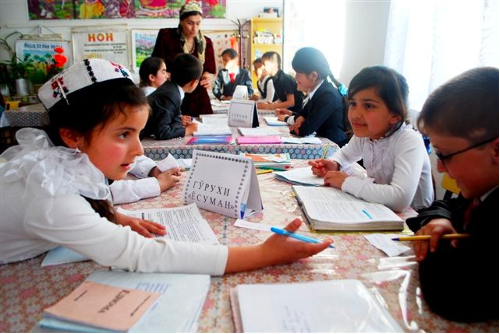 School children are seated around two large tables with school papers spread out. The children interact with each other while a teacher stands near one of the tables.