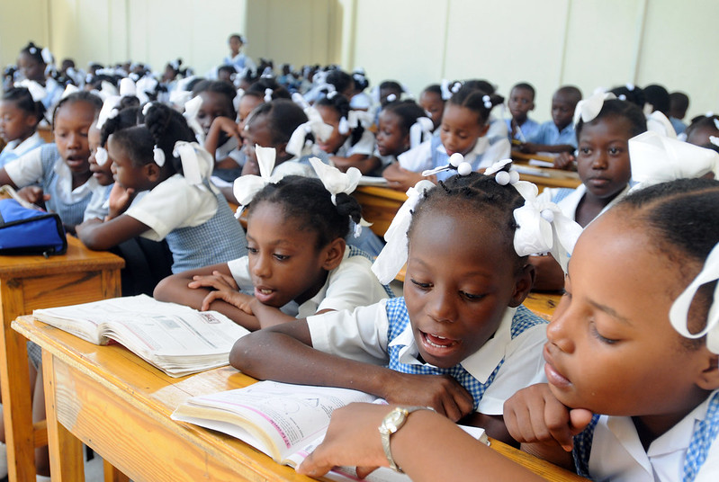 A large classroom of children sitting at shared tables. Many are sharing a textbook with the child next to them. All the children have blue and white shirts. Many of the girls also have white ribbons in their hair.